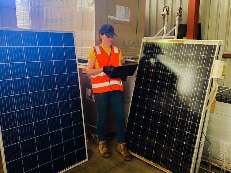 Mujer joven en alta visibilidad lleva piezas fotovoltaicas mientras pasa junto a dos viejos paneles solares