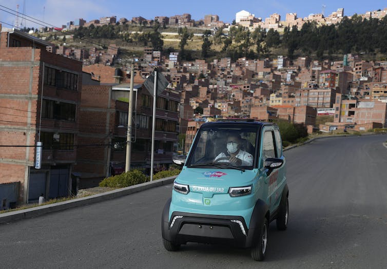 Un auto diminuto lo suficientemente grande para una persona, sin asientos para pasajeros, conduce por una calle de edificios de ladrillo.  Quantum Motors, su fabricante, tiene su sede en Bolivia.