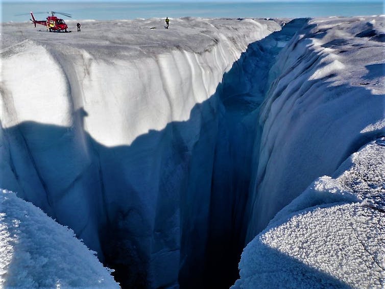 Un helicóptero sentado sobre la capa de hielo se ve diminuto al lado del enorme moulin, donde una corriente de agua derretida se vierte en la capa de hielo.