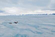 Vista aérea del océano cubierto de hielo con una hilera de picos montañosos a lo lejos. Un pequeño grupo de viajeros se reunió cerca de cañones de nieve y trineos de madera.