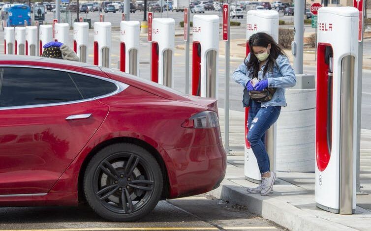 Una mujer estaba parada detrás de su auto eléctrico Tesla rojo, metiendo la mano en el bolsillo trasero, lista para enchufarlo en uno de los muchos puntos de carga.