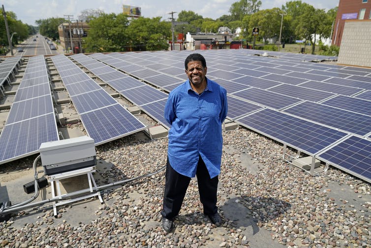 Un hombre parado en un techo con paneles solares y una comunidad al fondo.