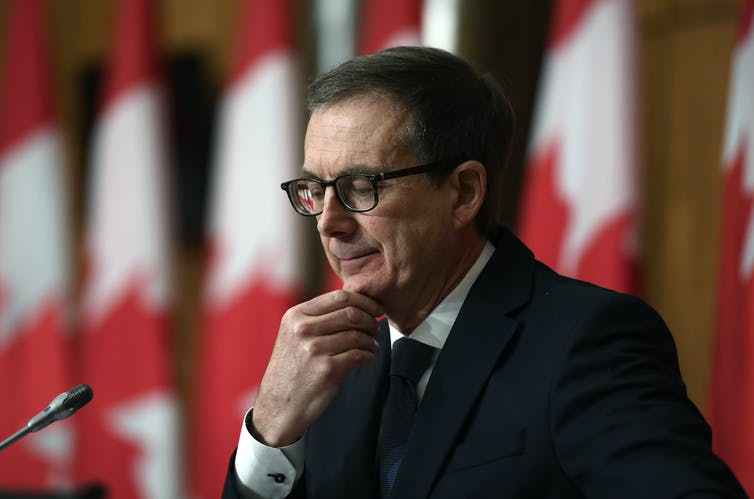 Un hombre con gafas en una conferencia de prensa con una fila de banderas canadienses detrás de él.