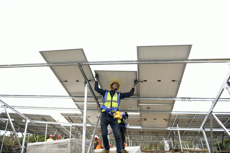 Un hombre con un casco se para debajo de tres grandes paneles solares.