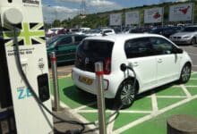 Un pequeño coche eléctrico blanco, estacionado en un estacionamiento verde, conectado a un punto de carga.