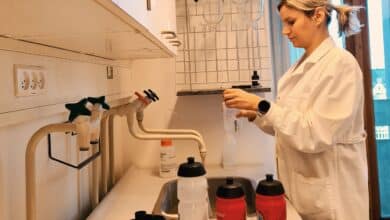 una mujer con una bata blanca se para en un fregadero de laboratorio junto a un mostrador que sostiene tres botellas deportivas de plástico