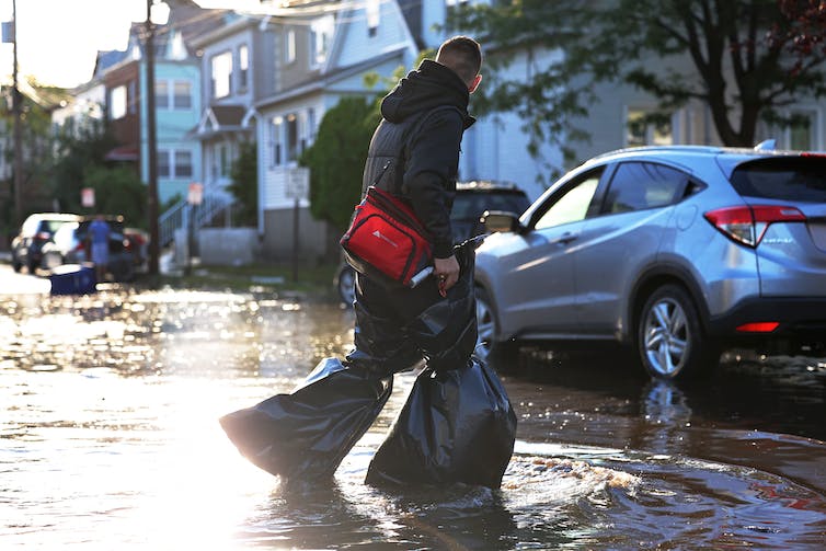 Una persona que usa bolsas de basura alrededor de cada pierna para mantenerlas secas cruza una calle inundada en Nueva Jersey. Un bote de basura volcado flota en el fondo.