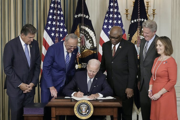 Biden se sienta en un escritorio firmando la legislación. Los senadores Joe Manchin (D-WV.) y Chuck Schumer (D-NY) y los representantes James Clyburn (D-SC), el representante Frank Pallone (D-NJ) y la representante Kathy Catsor (D-FL) revisan su hombro