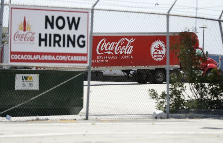 Un camión con remolque retrocede hacia un muelle de carga en Coca-Cola Beverages Florida más allá de un cartel de Now Hiring.
