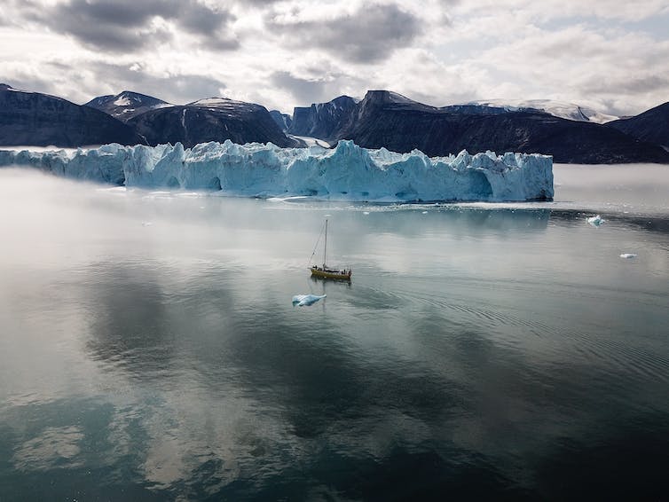 Un gran velero con un iceberg aún más grande detrás y un glaciar en la distancia.