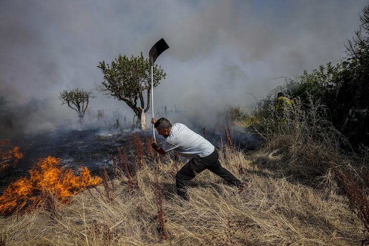 Un hombre golpea la hierba en llamas con una pala. El paisaje ennegrecido por el fuego está en el fondo.