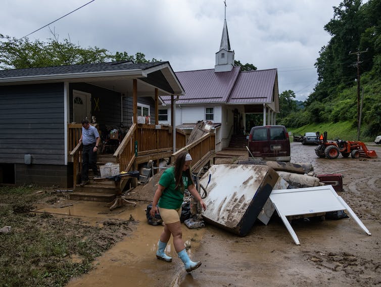 Una niña con botas de lluvia camina por un patio lleno de barro. Colchones dañados y otras pertenencias de una casa inundada se apilan cerca.
