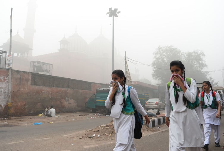 Tres niñas con uniformes escolares blancos caminan por una calle llena de smog con pañuelos sobre la nariz.