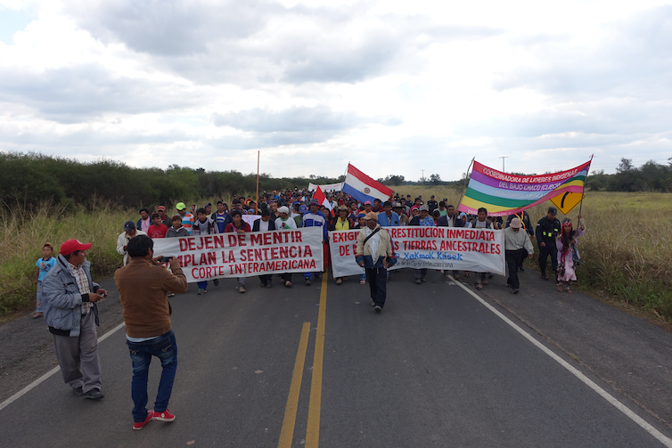 La gente camina por una carretera con pancartas que exigen que el Estado les devuelva sus tierras ancestrales.