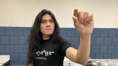 un joven con cabello largo y negro que usa una camisa negra se para en un laboratorio y sostiene un fósil tardígrado entre sus dedos