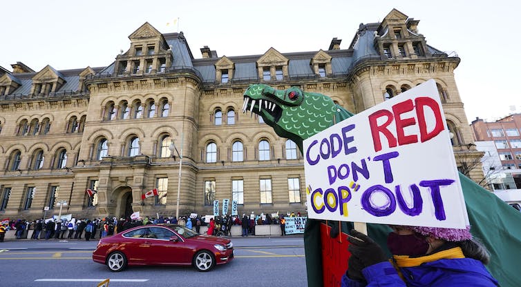 Una persona que lleva un cartel que dice código rojo, no se escabulle frente a un edificio de piedra con otros manifestantes.
