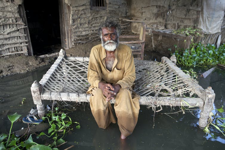 Un hombre se sienta en un banco frente a la puerta de su casa, rodeado de agua hasta las espinillas.