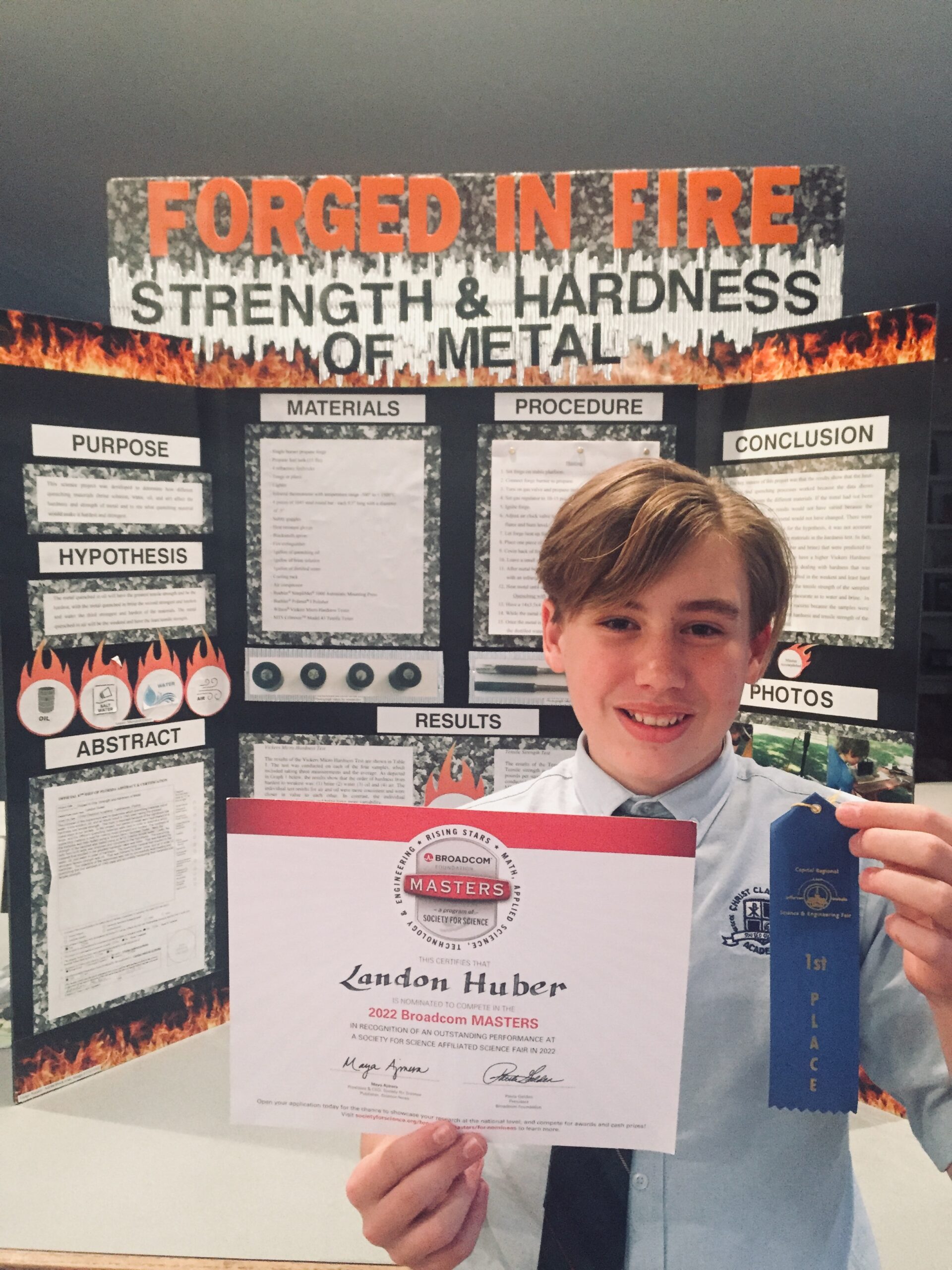 un chico rubio se para frente a un cartel de proyecto titulado 'Forjado en fuego' y sostiene una cinta azul y un certificado