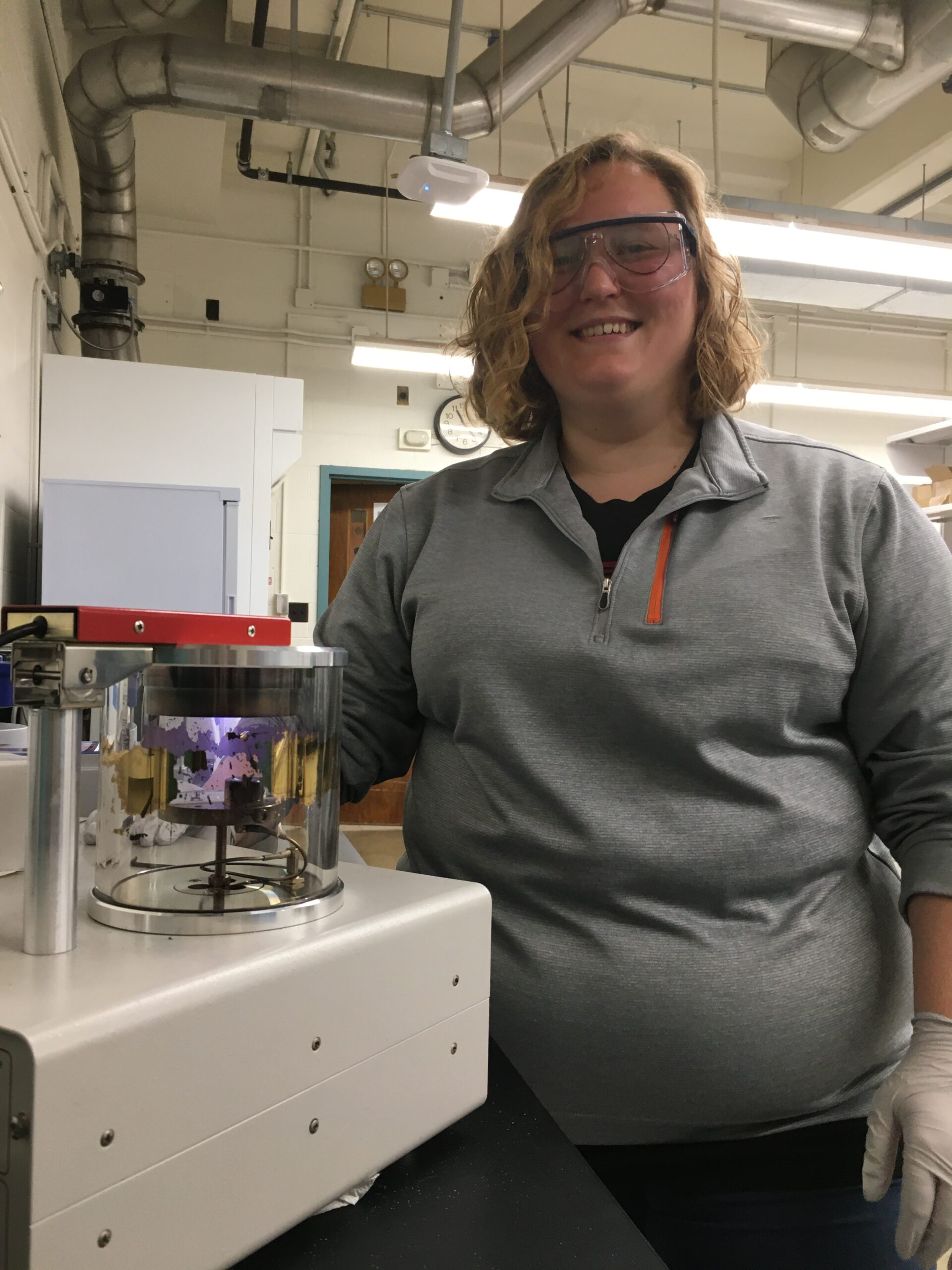una joven rubia con gafas se encuentra junto a una pieza de maquinaria en un laboratorio