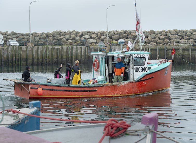 La gente se para en la cubierta de un pequeño barco de pesca que flota en el puerto de un cuerpo de agua.