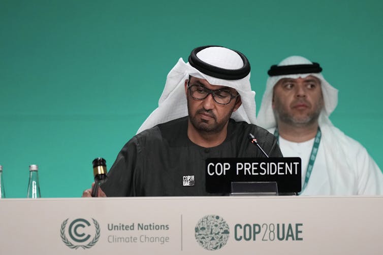 Un hombre del sur de Asia con gafas y una kufiyah blanca golpea un pequeño mazo sobre una mesa con el logo de la COP impreso en el frente. Una placa con su nombre frente a él dice PRESIDENTE DE LA COP.