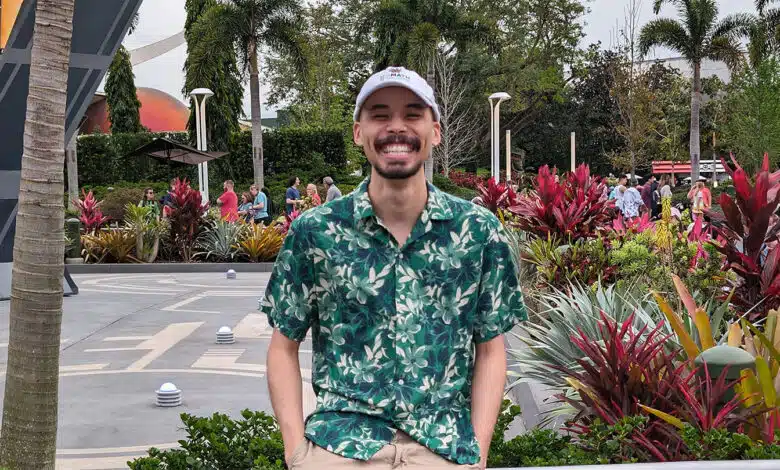 Computer Scientist Niall Williams stands in front of metal handrail. He's has a black moustache and beard. He's wearing a powder blue baseball cap and a button down shirt with tree designs on it. A concrete courtyard and palm trees are in the background.