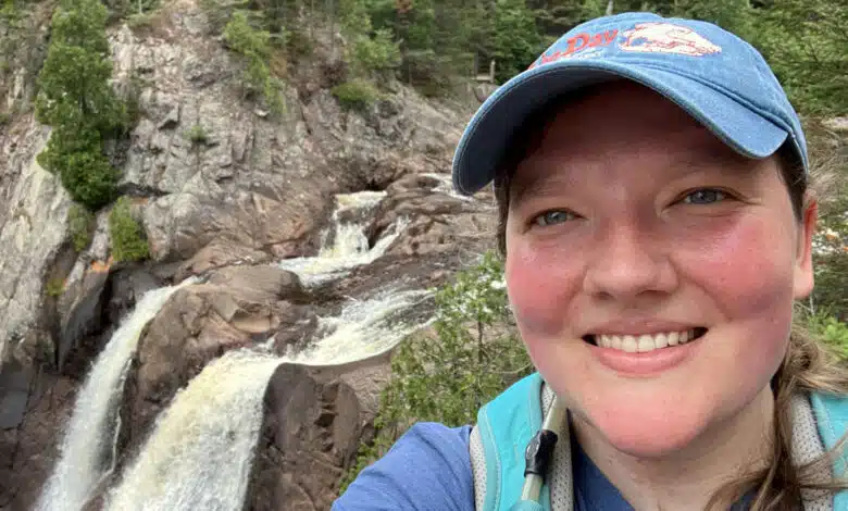 Anne Kort takes a selfie in front of High Falls at Tettegouche State Park in northern Minnesota. She's wearing a blue baseball cap and a teal backpack.