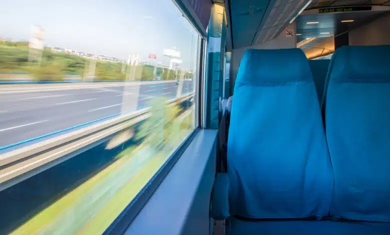 Two plush blue seats are against the window of a dimly lit train car, while outside the view of a highway and blue sky whizzes by so fast it's blurry
